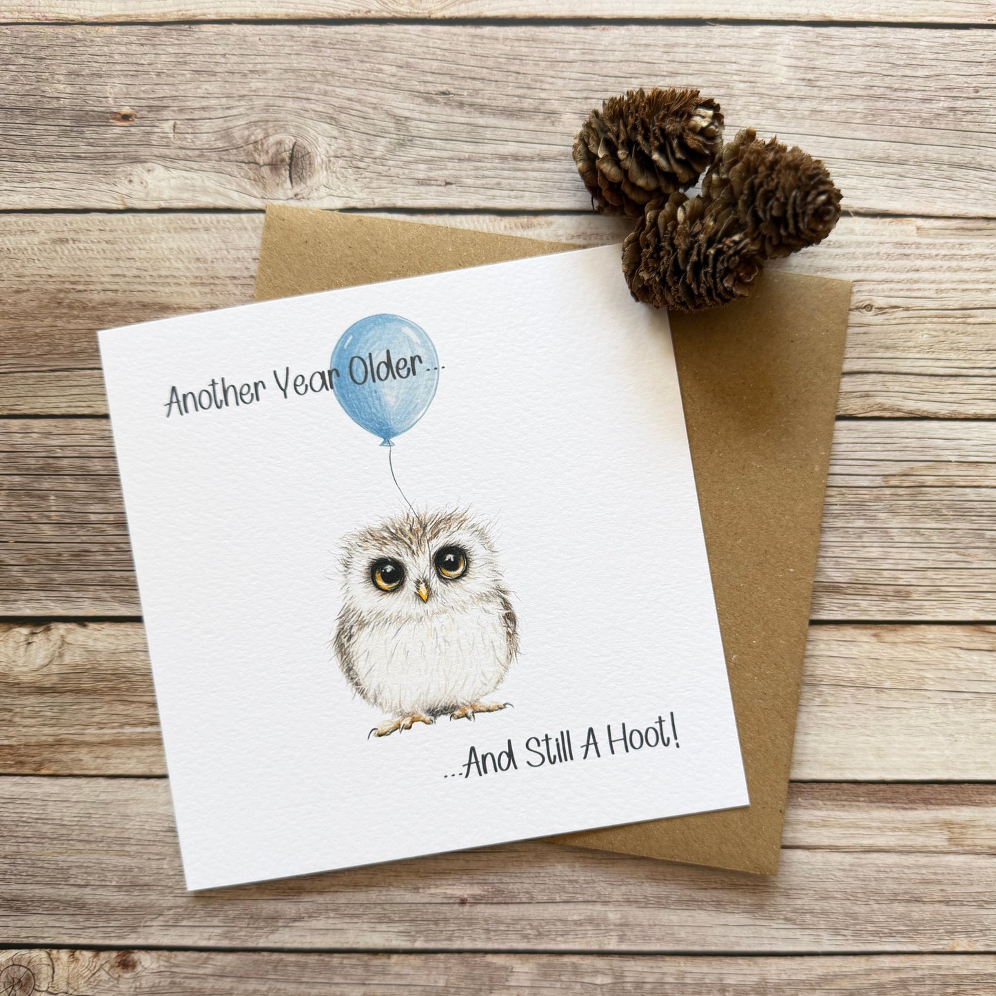 A square white greeting card with a watercolor-style owl holding a blue balloon, placed on top of a brown kraft envelope on a rustic wood plank surface. The card reads, 'Another Vean Older.