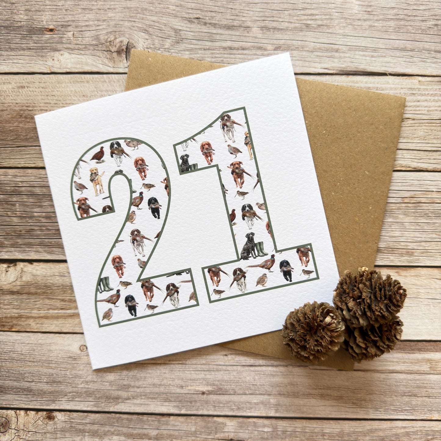 A square white greeting card with the number 21 filled with small illustrated figures, placed on top of a brown kraft envelope on a rustic wooden surface, with two pinecones nearby.