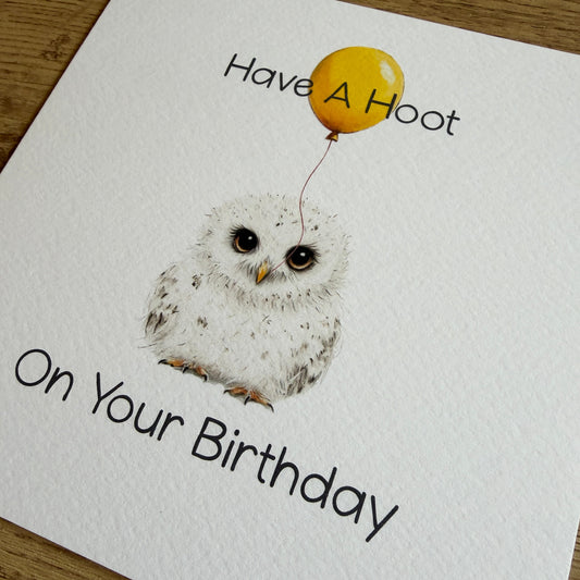 A square white greeting card with a watercolor-style baby owl illustration and the text 'Have A Hoot On Your Birthday,' photographed on a wooden surface.