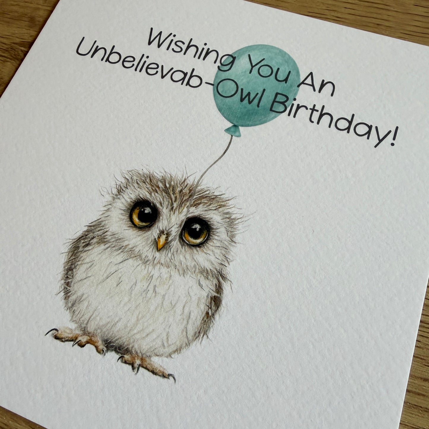 A square white greeting card featuring a hand-drawn owl with large eyes and a small teal balloon, with the text 'Wishing You An Unbelievab-Owl Birthday!' printed on it, photographed on a wooden surface.