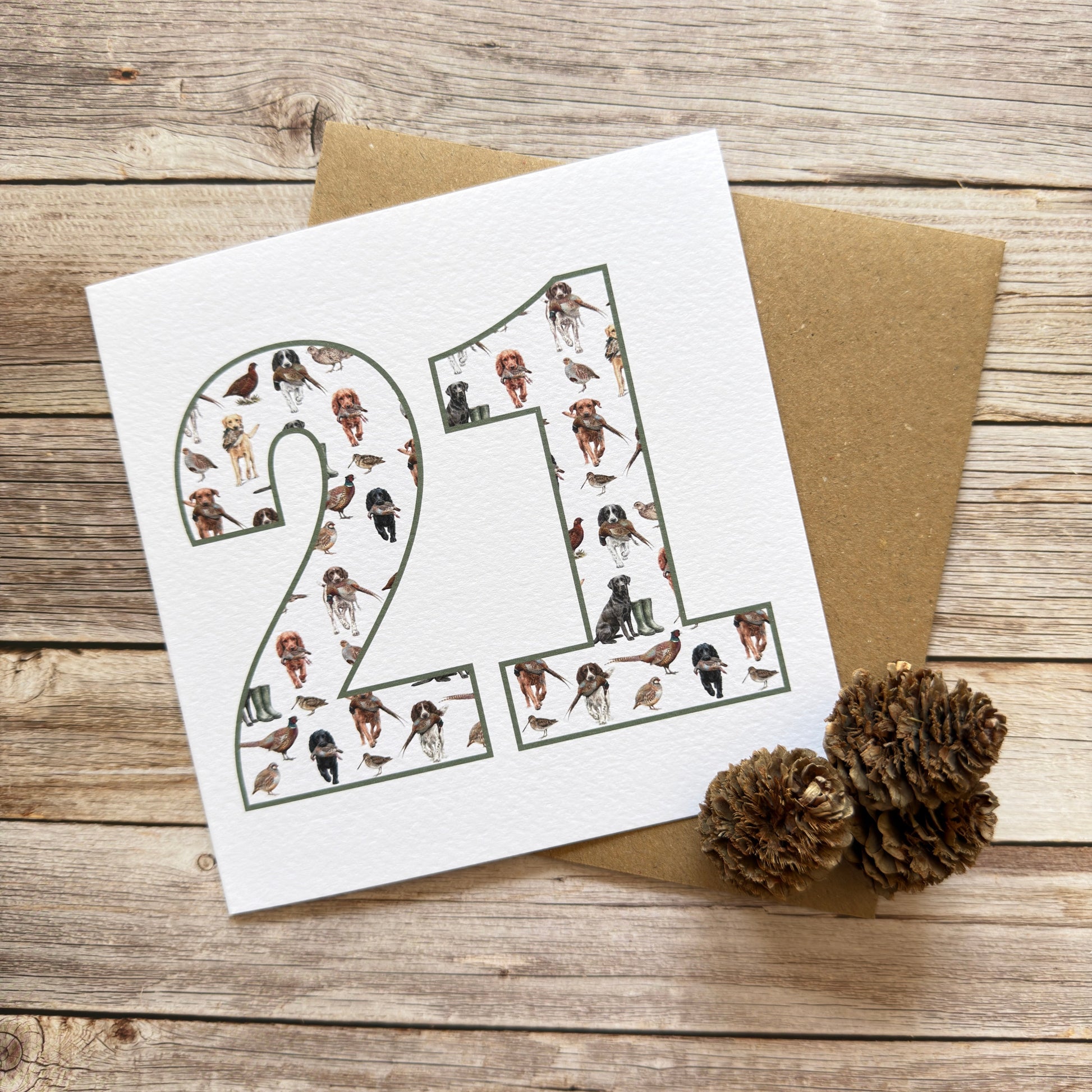 A square white greeting card with the number 21 filled with small illustrated figures, placed on top of a brown kraft envelope on a rustic wooden surface, with two pinecones nearby.