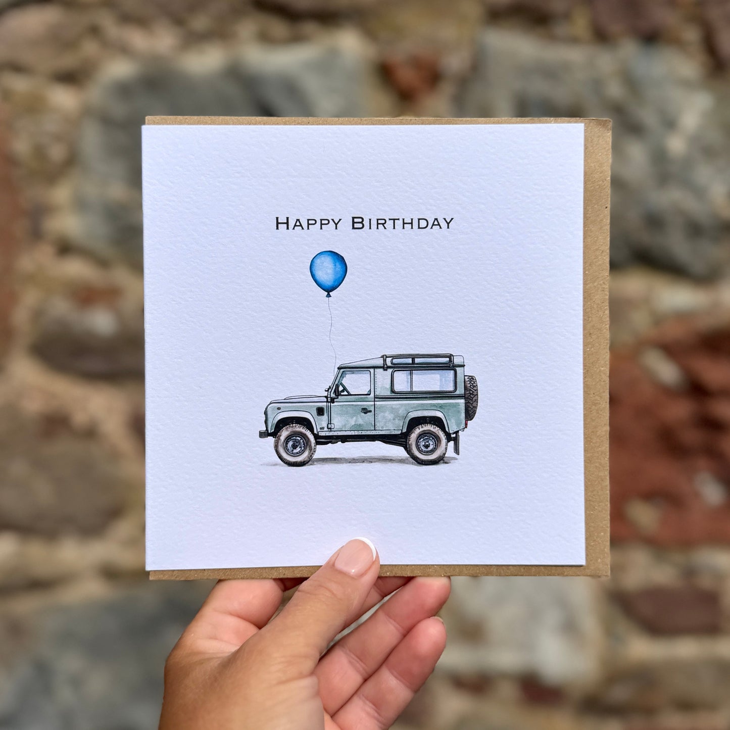 A hand holds a square birthday card in front of a blurred stone wall. The card has a white front with the text 'HAPPY BIRTHDAY' above a watercolor-style illustration of a light blue vehicle holding a blue balloon, with a brown kraft paper border.