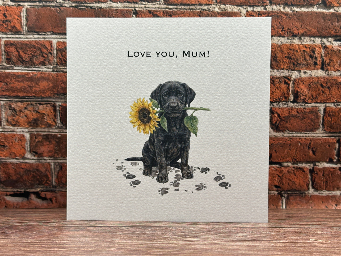 Drawing of a Black Labrador puppy sitting with muddy paw prints, holding a large yellow sunflower in its mouth with text reading Love You, Mum.