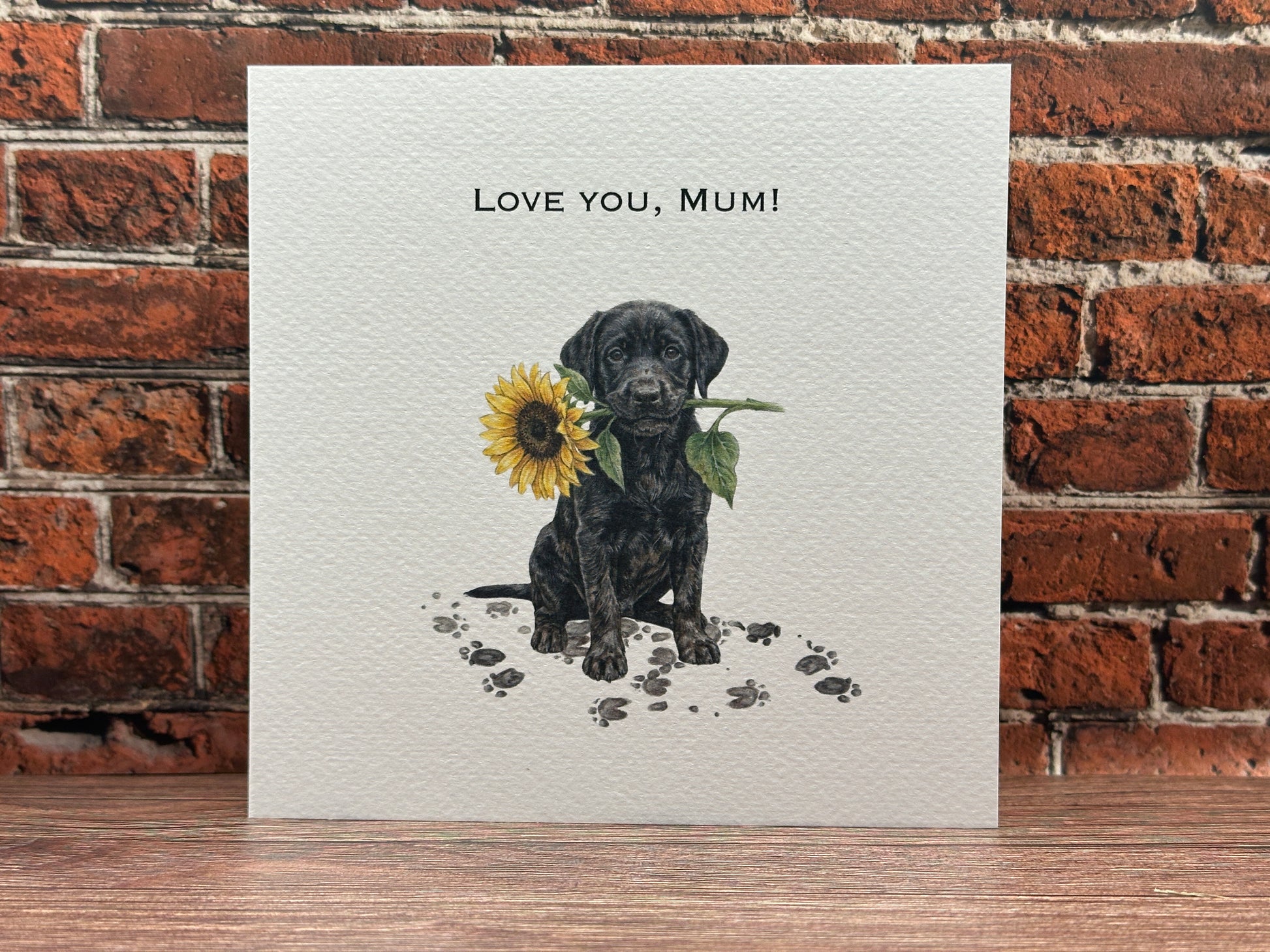 Drawing of a Black Labrador puppy sitting with muddy paw prints, holding a large yellow sunflower in its mouth with text reading Love You, Mum.
