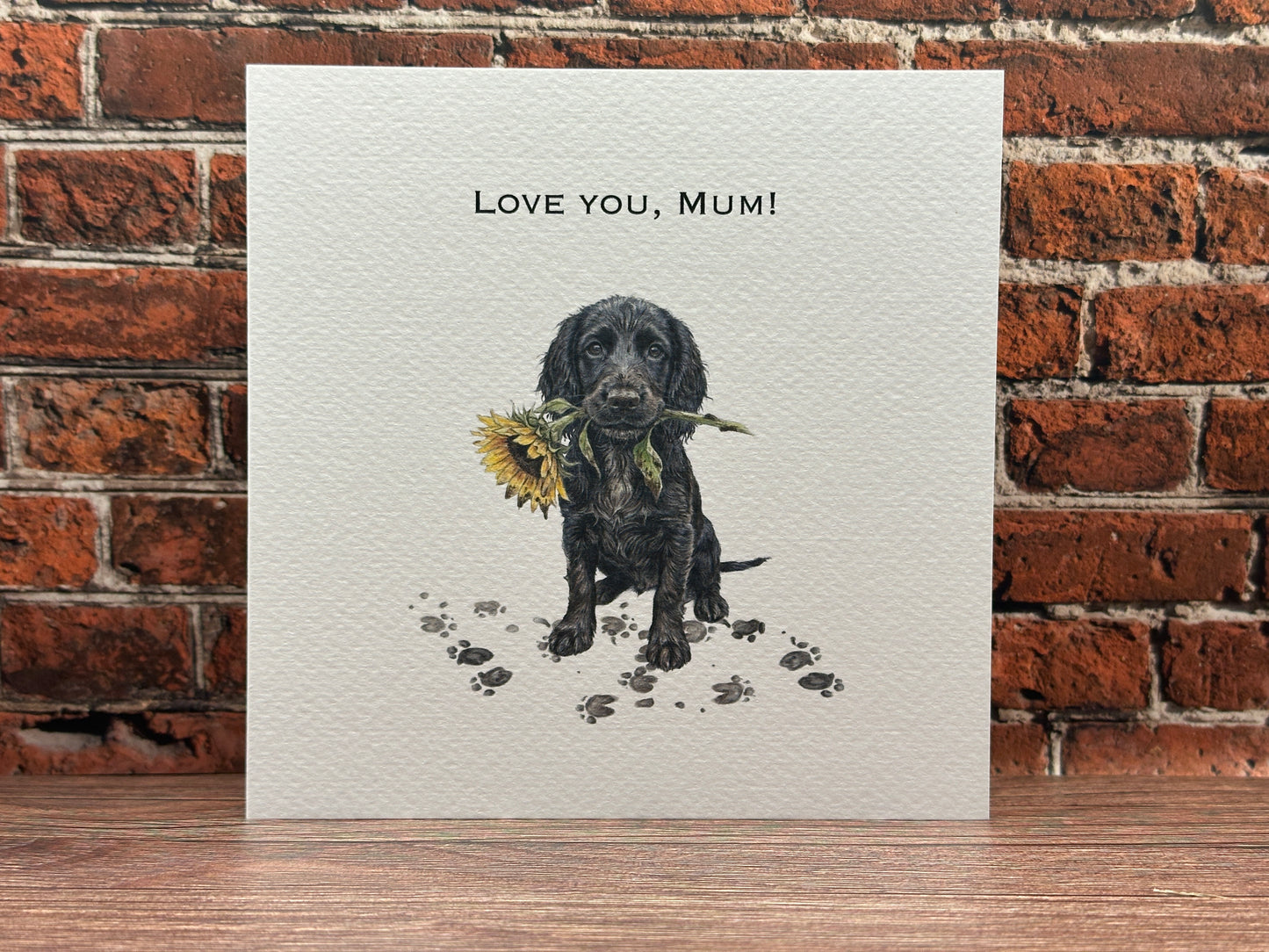 Illustration of a Black Cocker Spaniel puppy sitting with muddy paw prints, holding a yellow sunflower in its mouth, with text reading Love You, Mum.