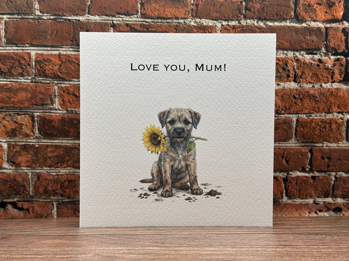 Illustration of a Border Terrier puppy sitting with muddy paw prints, holding a yellow sunflower in its mouth, with text reading Love You, Mum.