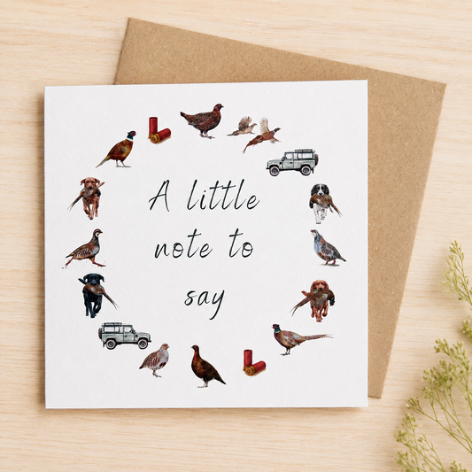 A square white greeting card with the phrase 'A little note to say' in black script, surrounded by small illustrated birds, rabbits, and vehicles, styled on a light wood surface with a brown envelope behind it.